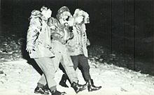 Black and white photograph of an airman in a bomber jacket being assisted to walk by two men
