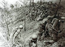 Two soldiers armed with a flame thrower are walking to the right with two soldiers armed with rifles. In the background a group of soldiers are resting over a desolate landscape