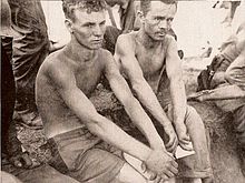 Two men without shirts on sit surrounded by soldiers