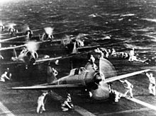 Planes on the deck of an aircraft carrier, with technical crews in white overalls attending the planes.