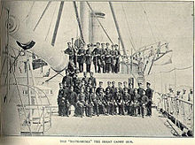 Large naval gun aboard a warship, with officier sitting on the deck under and above the gun.